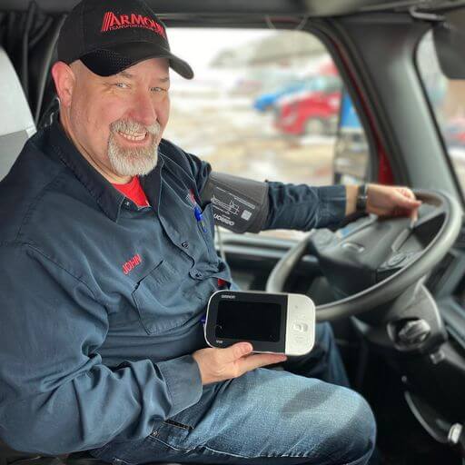 A trucker seated inside his truck is holding up a health tracking device.